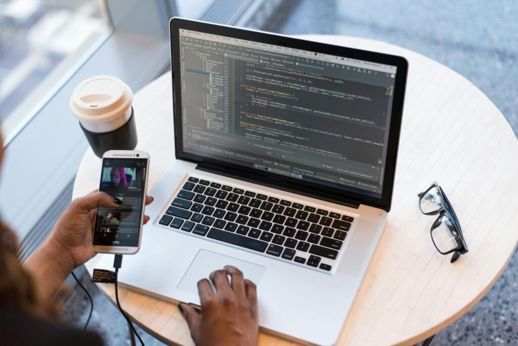 A programmer manages code on a laptop while multitasking with a smartphone, illustrating modern tech workspace.